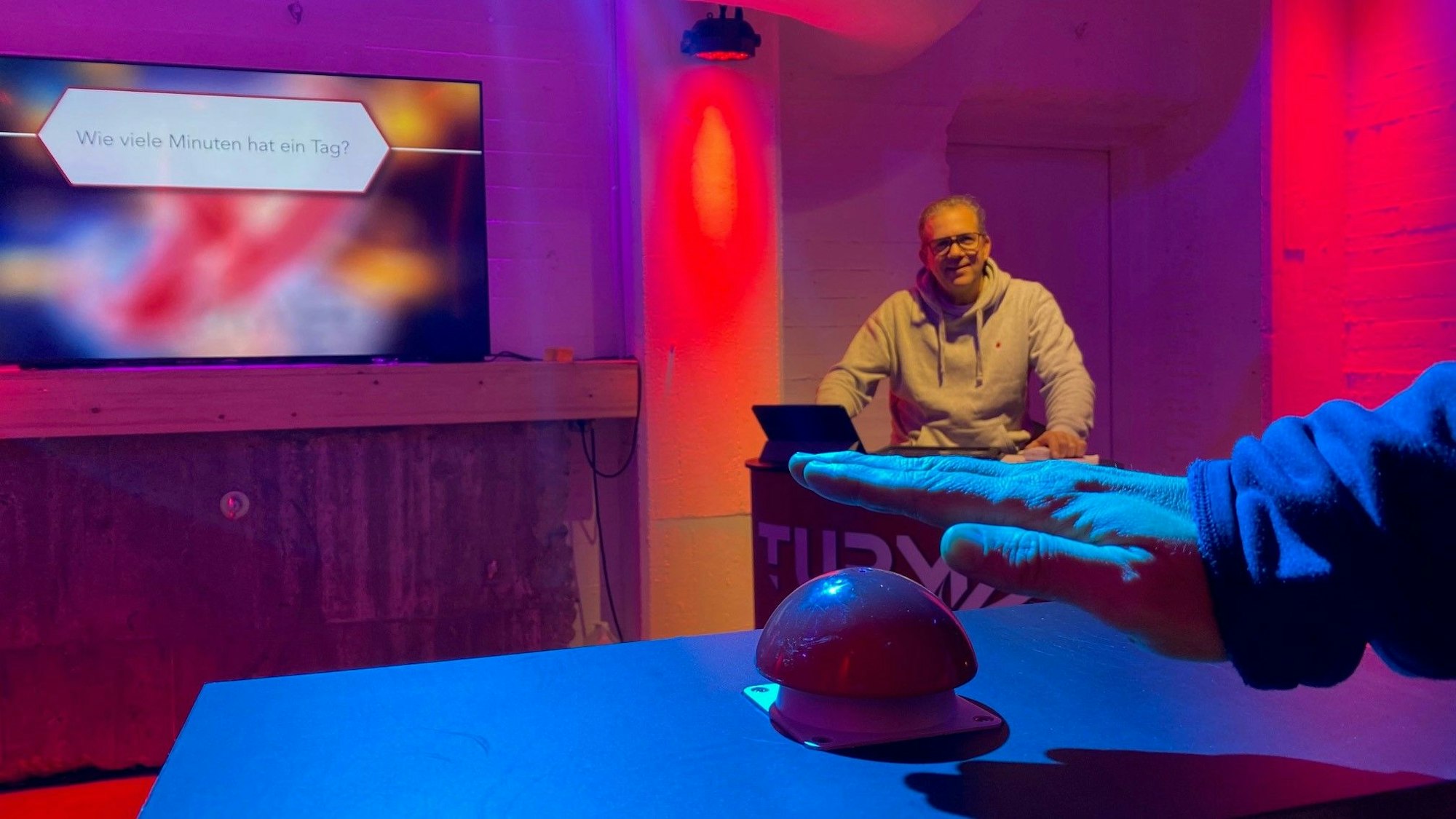 Zu sehen ist ein Buzzer-Knopf und im Hintergrund ein Monitor.