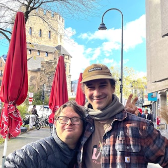 Ein kleiner Mann mit Brille wird von einem großen mit Kappe in den Arm genommen.
