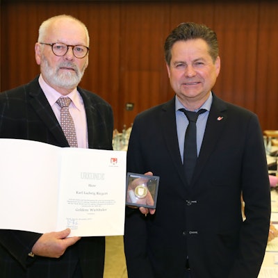 Zwei Männer, der linke hält eine Urkunde und eine Münze.