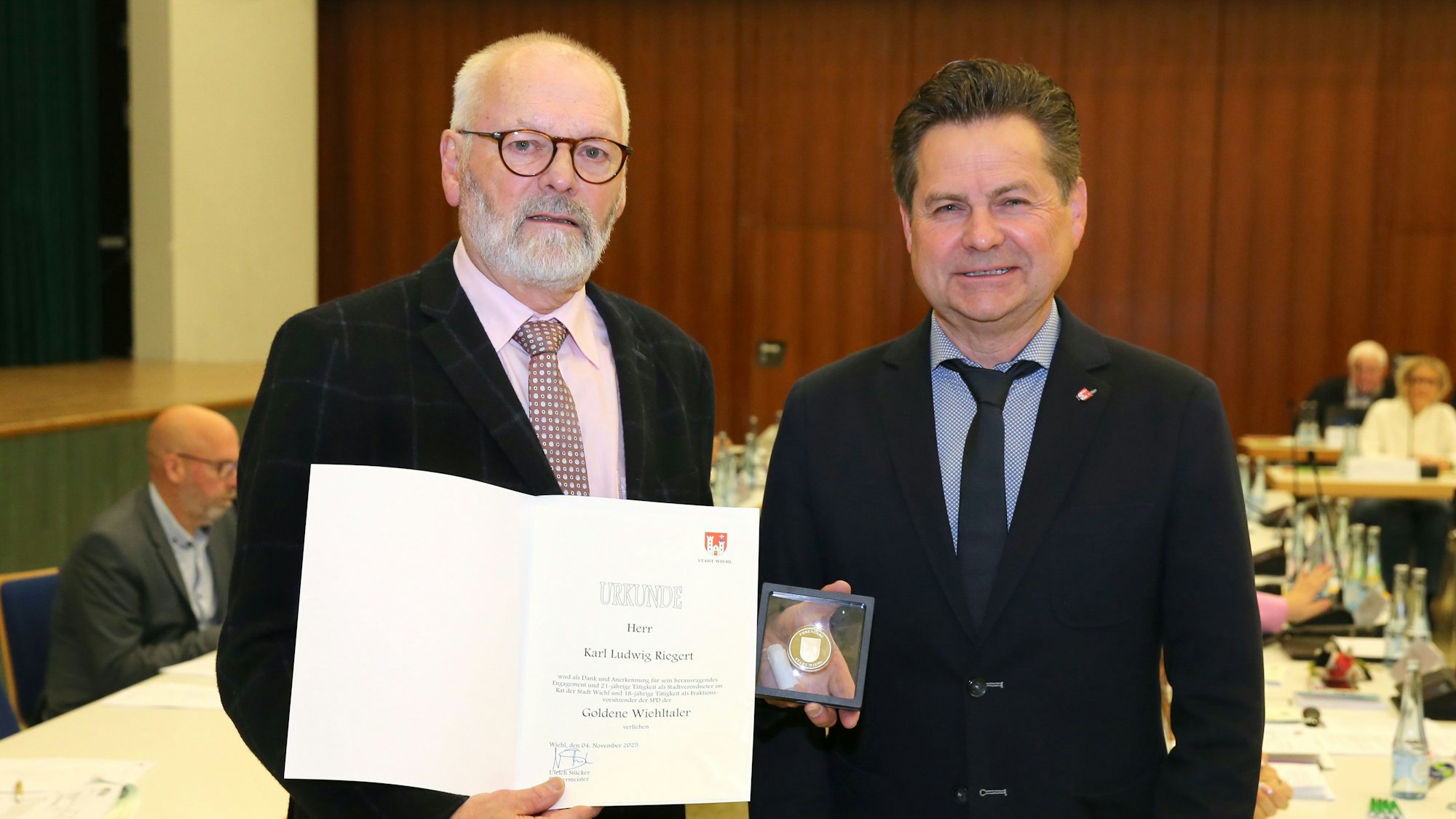Zwei Männer, der linke hält eine Urkunde und eine Münze.