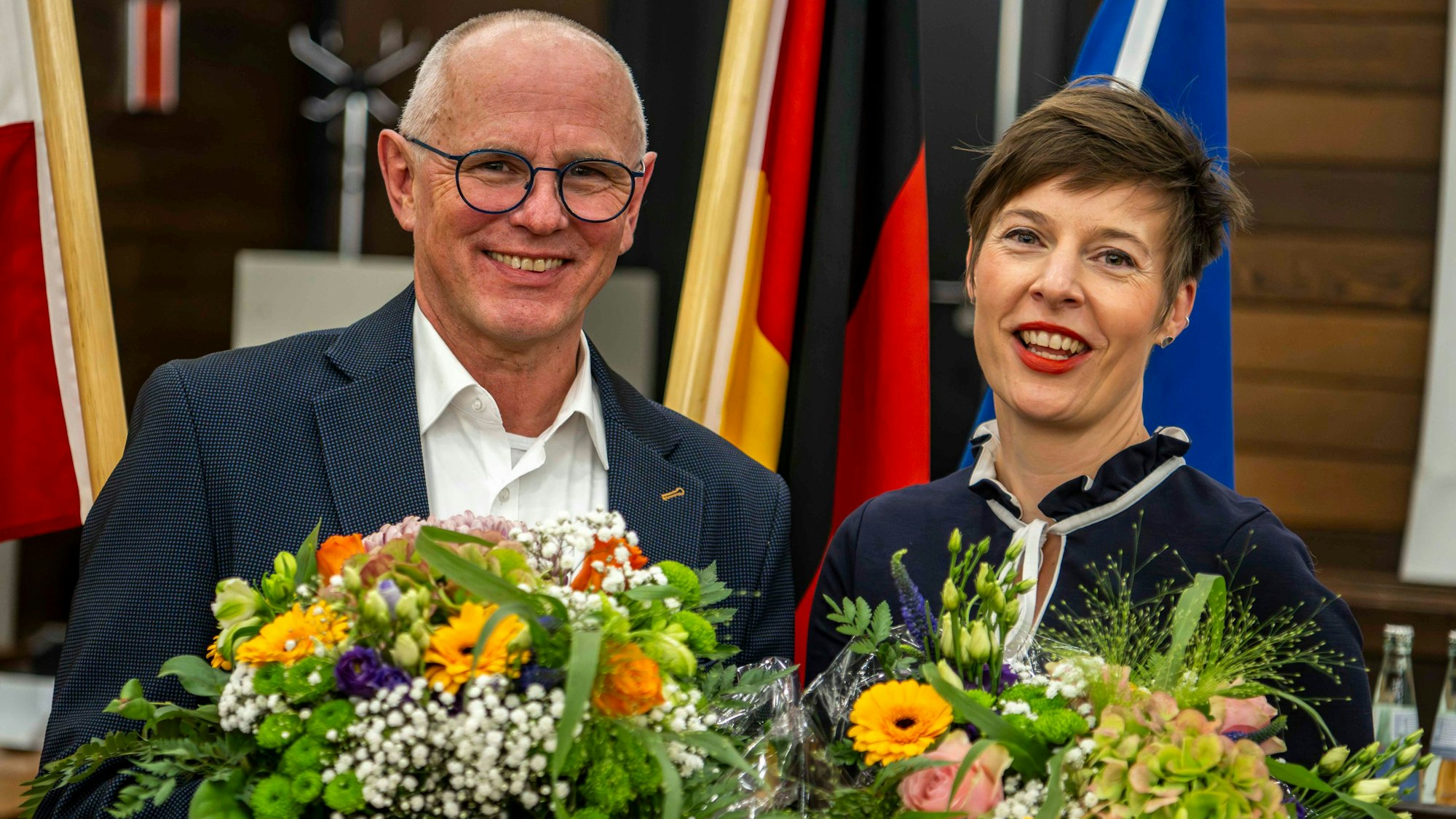 Ralf Claßen und Annegret Lewak stehen im Sitzungssaal des Kreishauses. Sie haben Blumensträuße in der Hand, im Hintergrund sind mehrere Fahnen zu sehen.
