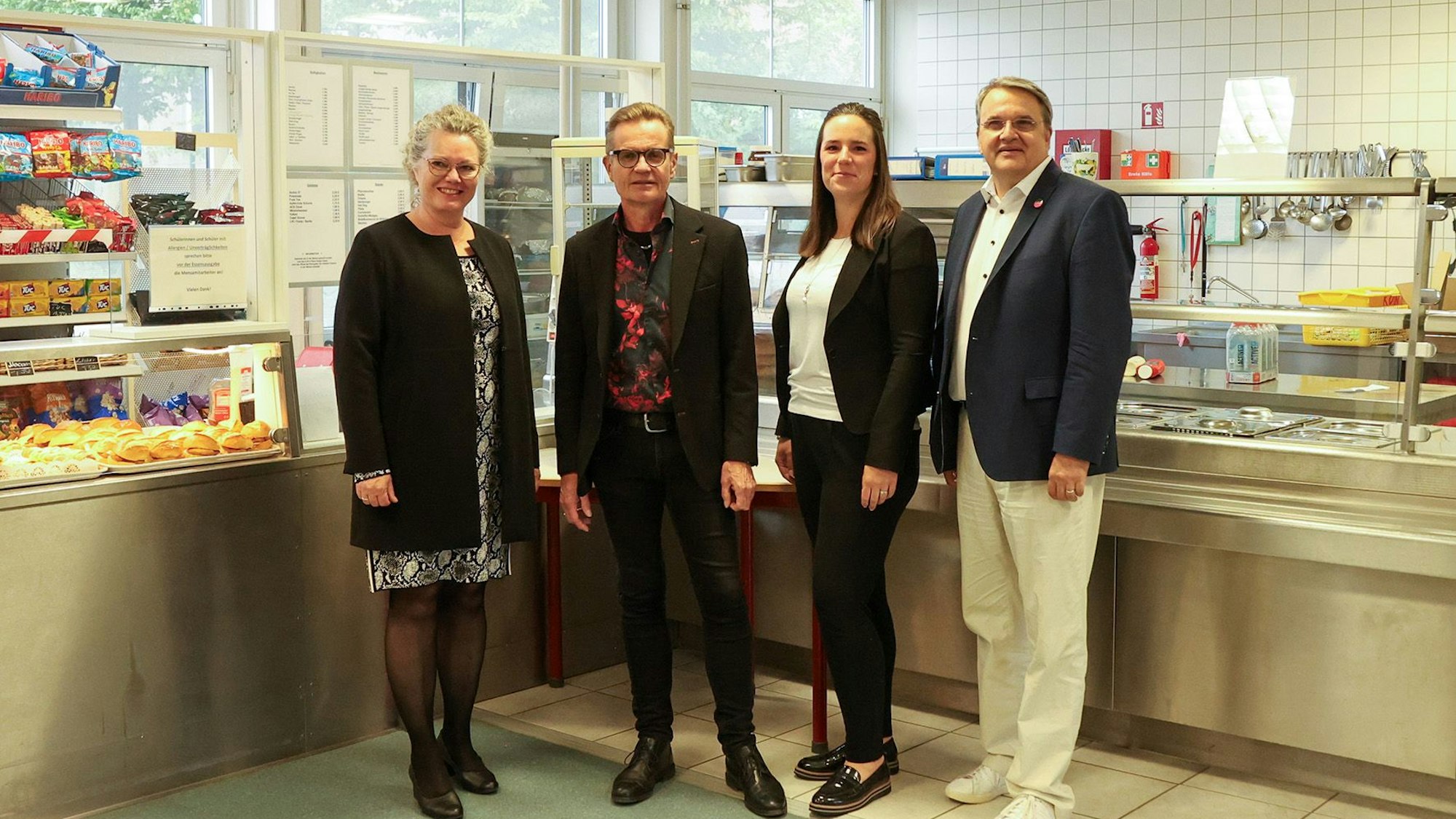 Die Gruppe steht in einer Schulcafeteria. In einem Regal befinden sich verschiedenen Snacks.