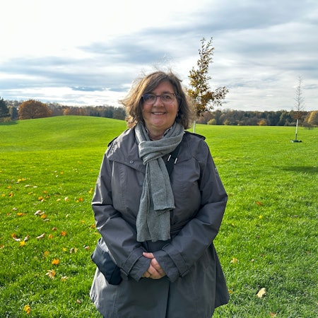 Ein Frau, die einen grauen Schal und Anorak trägt, steht auf einer weiten, hügeligen Wiese.
