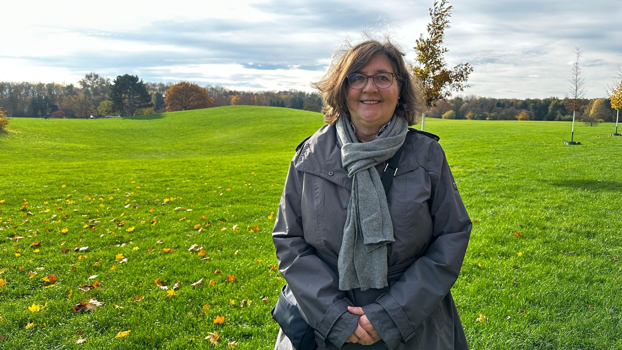 Ein Frau, die einen grauen Schal und Anorak trägt, steht auf einer weiten, hügeligen Wiese.
