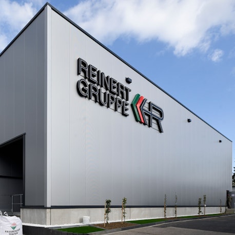 Zu sehen ist eine graue Lagerhalle vor blauem Himmel mit der Aufschrift „Reinert Gruppe“.