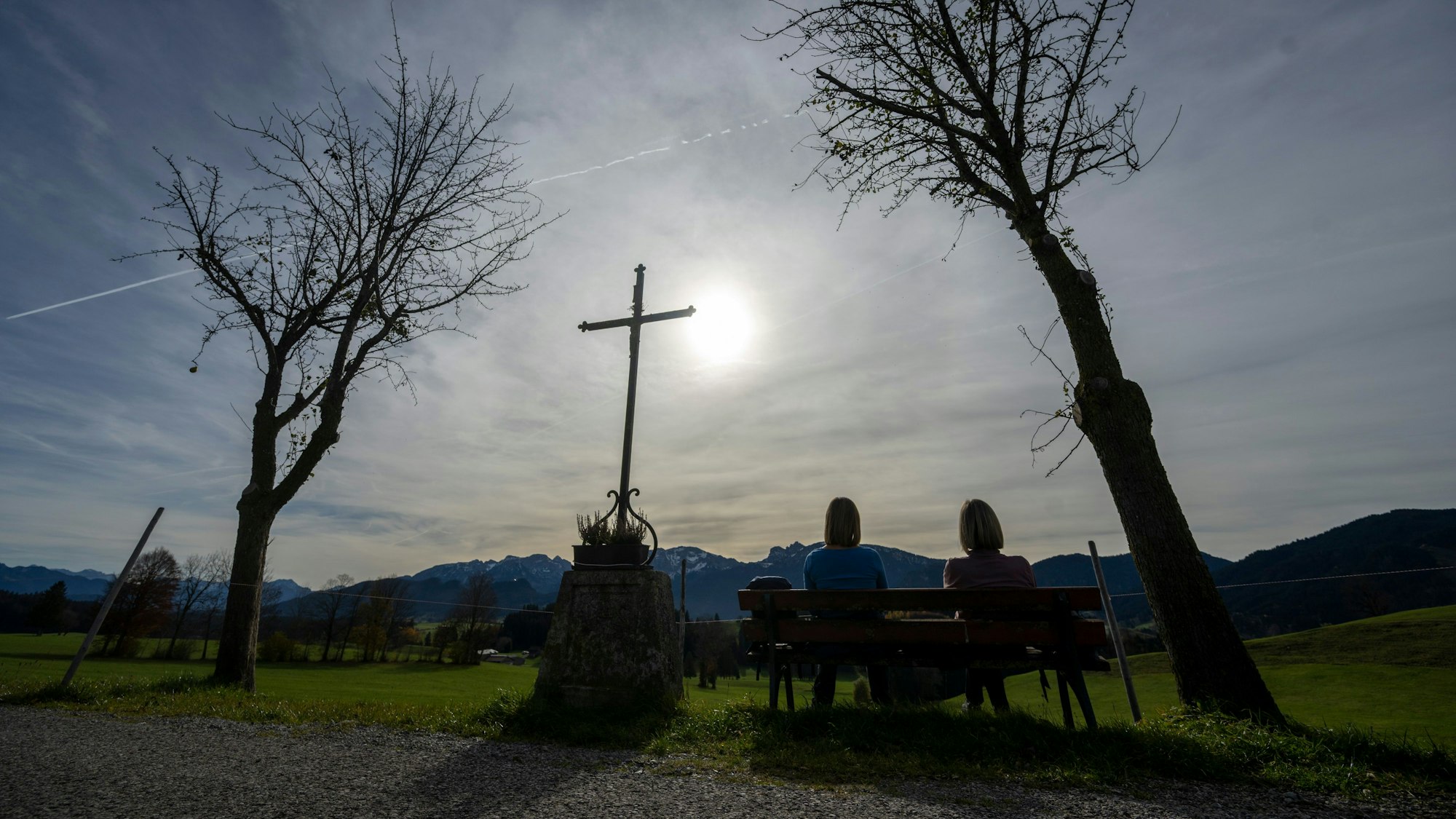 wei Frauen genießen auf eine Bank neben einem Wegkreuz die Herbstsonne.