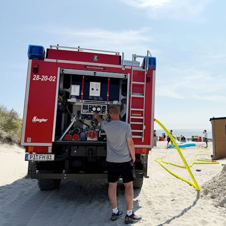 Ein Mann am Feuerwehrauto, ein Schlauch führt zum Strand.