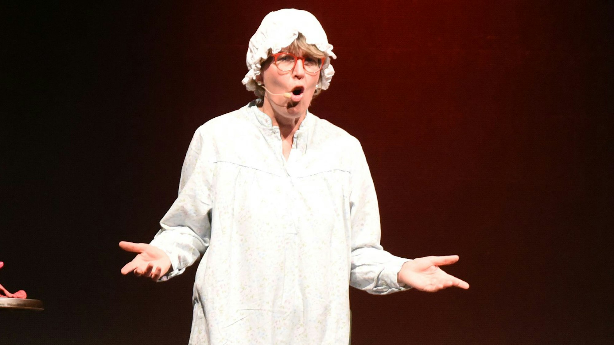 In Nachthemd, Gummistiefeln und mit Nachthaube auf dem Kopf stehr Katrin Iskam auf einer Bühne.