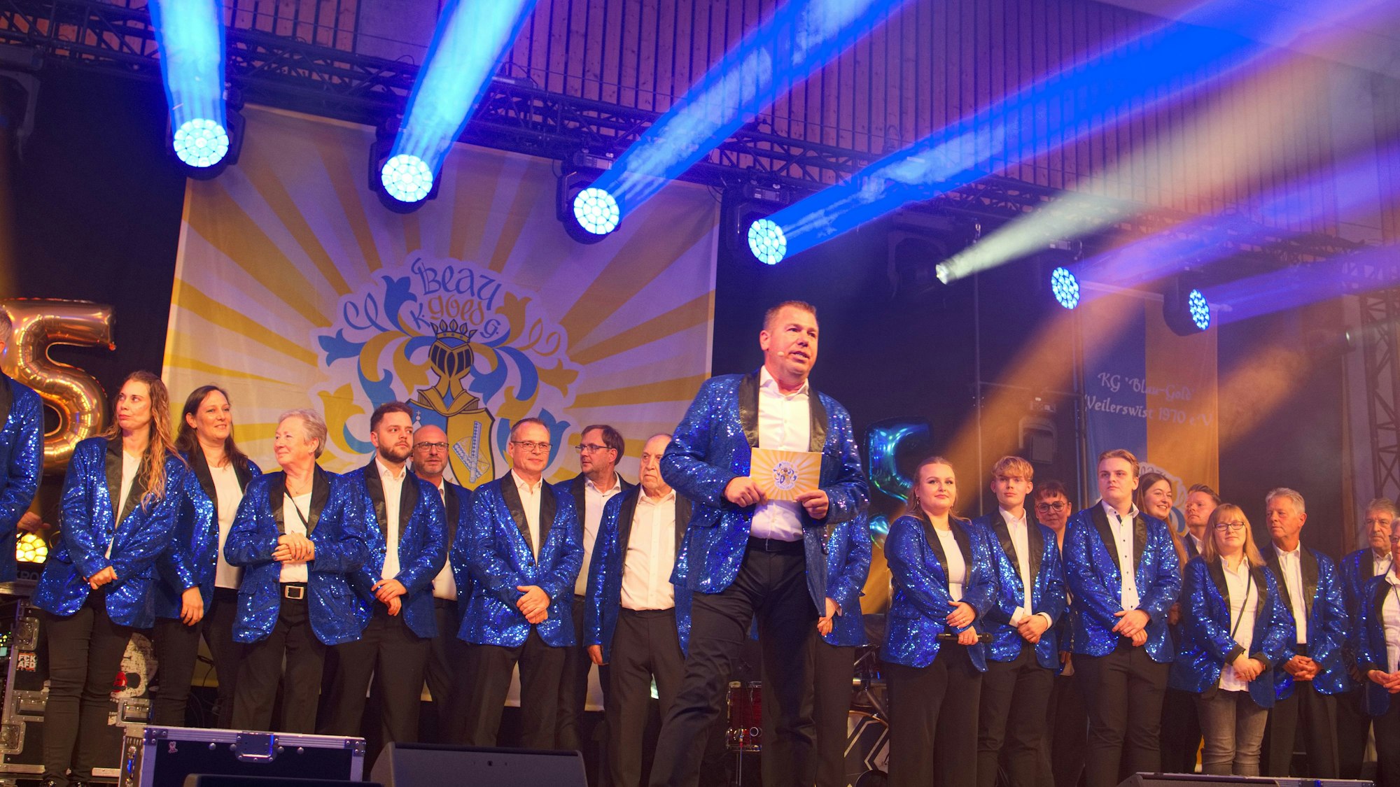 Männer und Frauen, einheitlich in blauen Jacketts und schwarzen Hosen, stehen nebeneinander auf der Bühne.