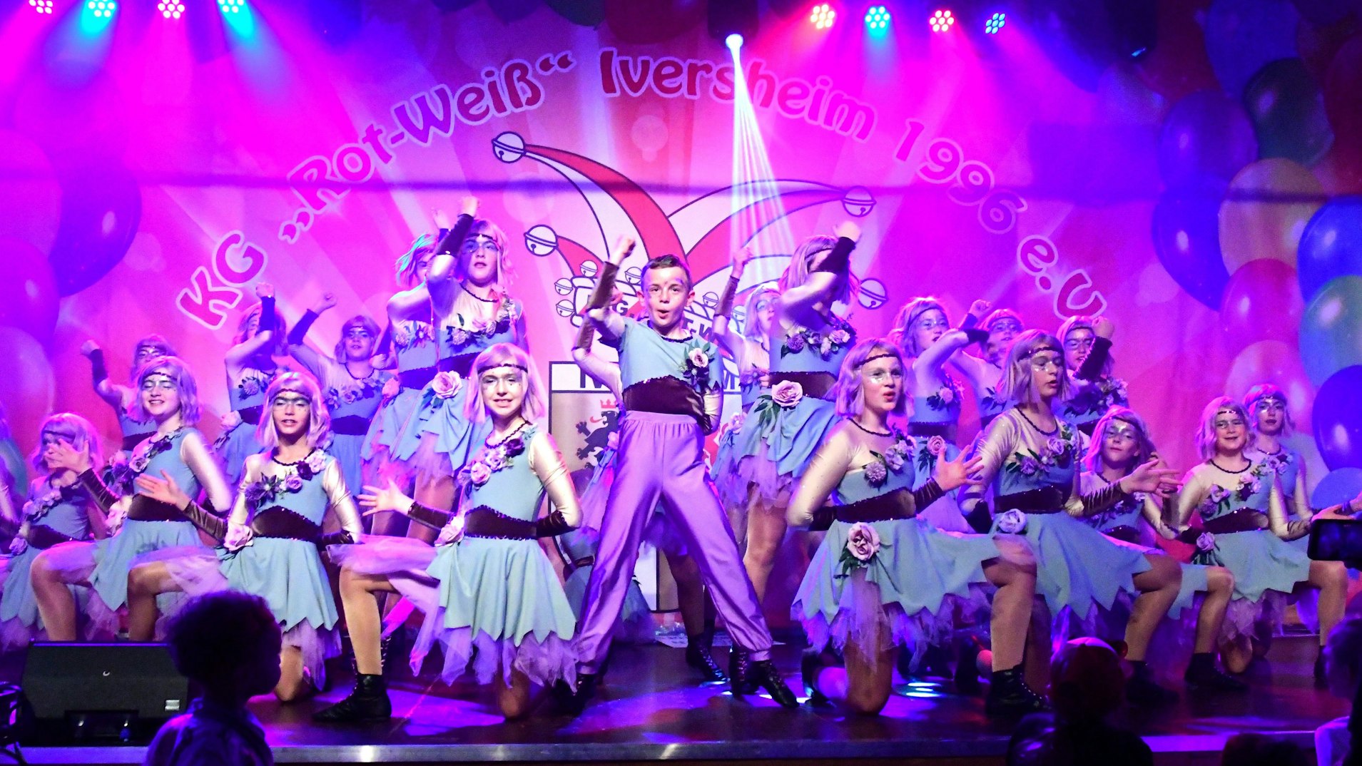 Kostümierte Mädchen knien um einen Jungen auf einer Karnevalsbühne.