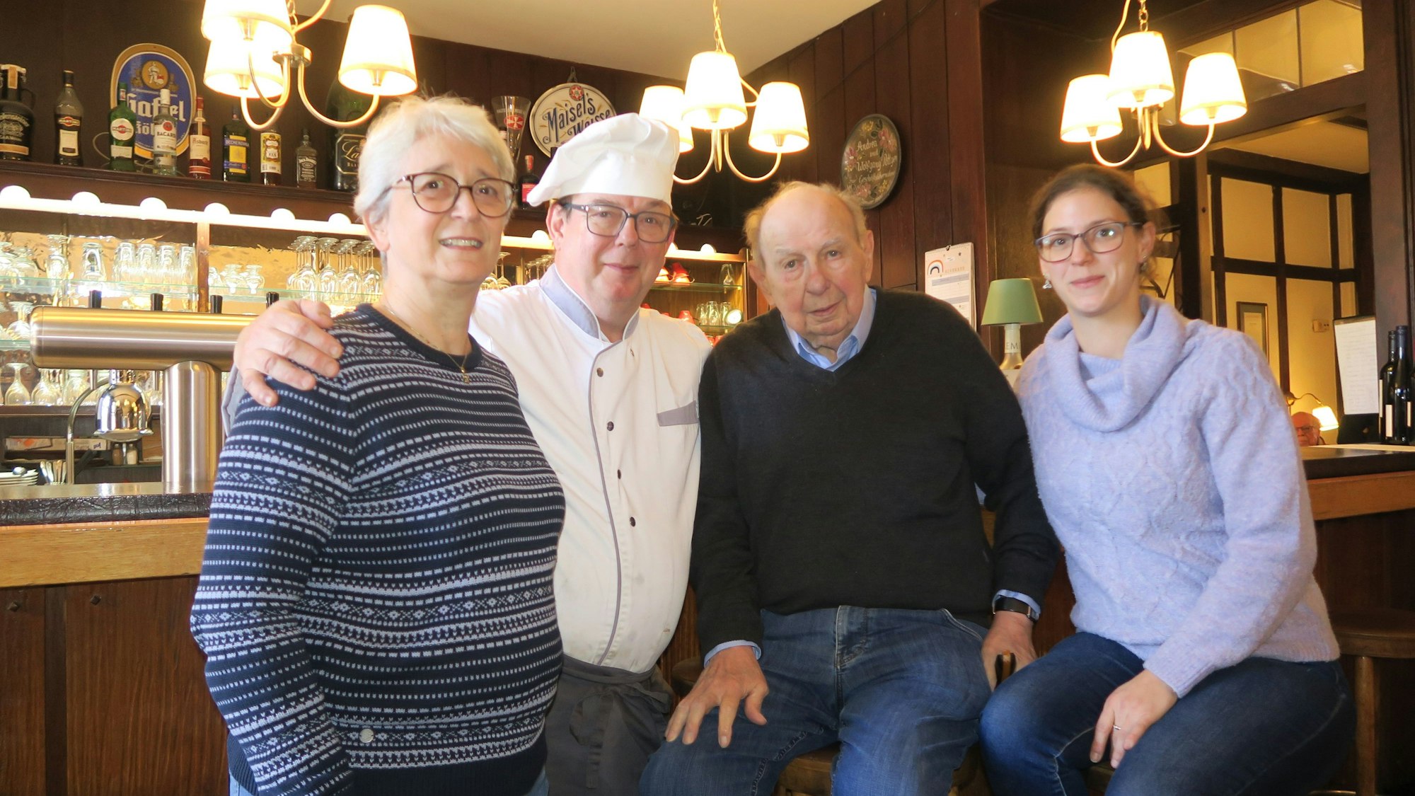 Zwei Frauen und zwei Männer stehen und sitzen auf Barhockern im Gastraum einer Wirtschaft.