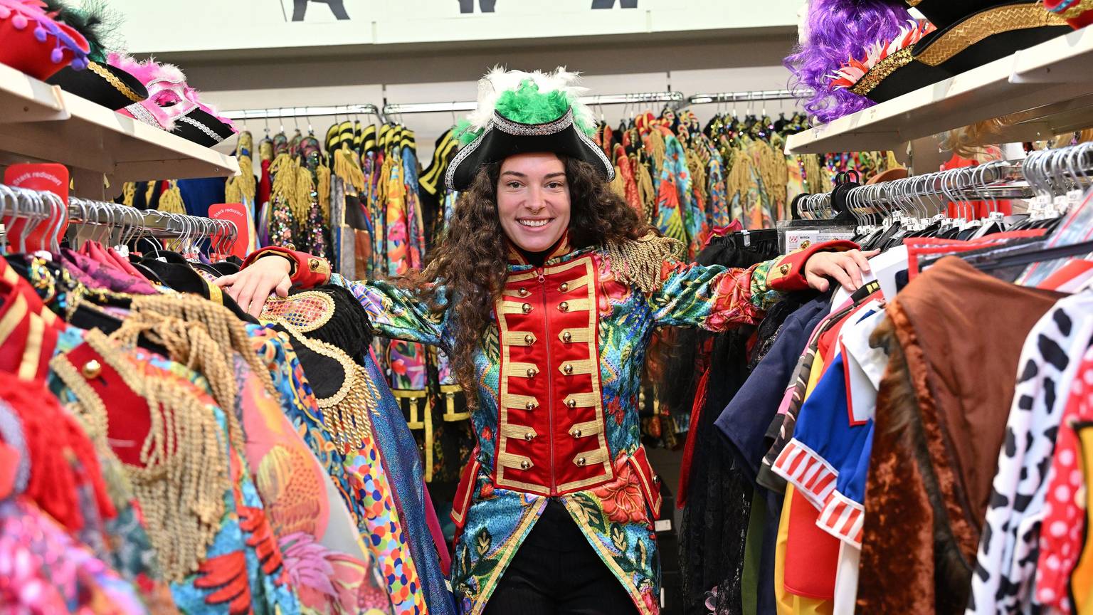 Eine Frau steht zwischen zwei Kleiderstangen an denen diverse Karnevalskostüme hängen. Sie trägt eine bunte Garde-Jacke und einen schwarzen Hut mit grünen Federn.
