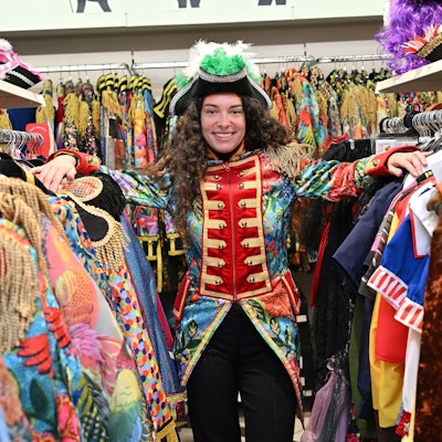 Eine Frau steht zwischen zwei Kleiderstangen an denen diverse Karnevalskostüme hängen. Sie trägt eine bunte Garde-Jacke und einen schwarzen Hut mit grünen Federn.