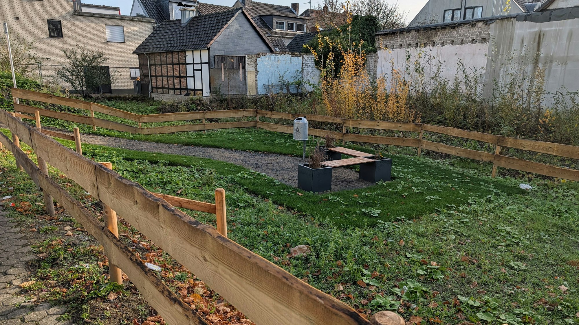 Das Foto zeigt die kleine, etwas verwildert wirkende Grünanlage hinter einem Bretterzaun.