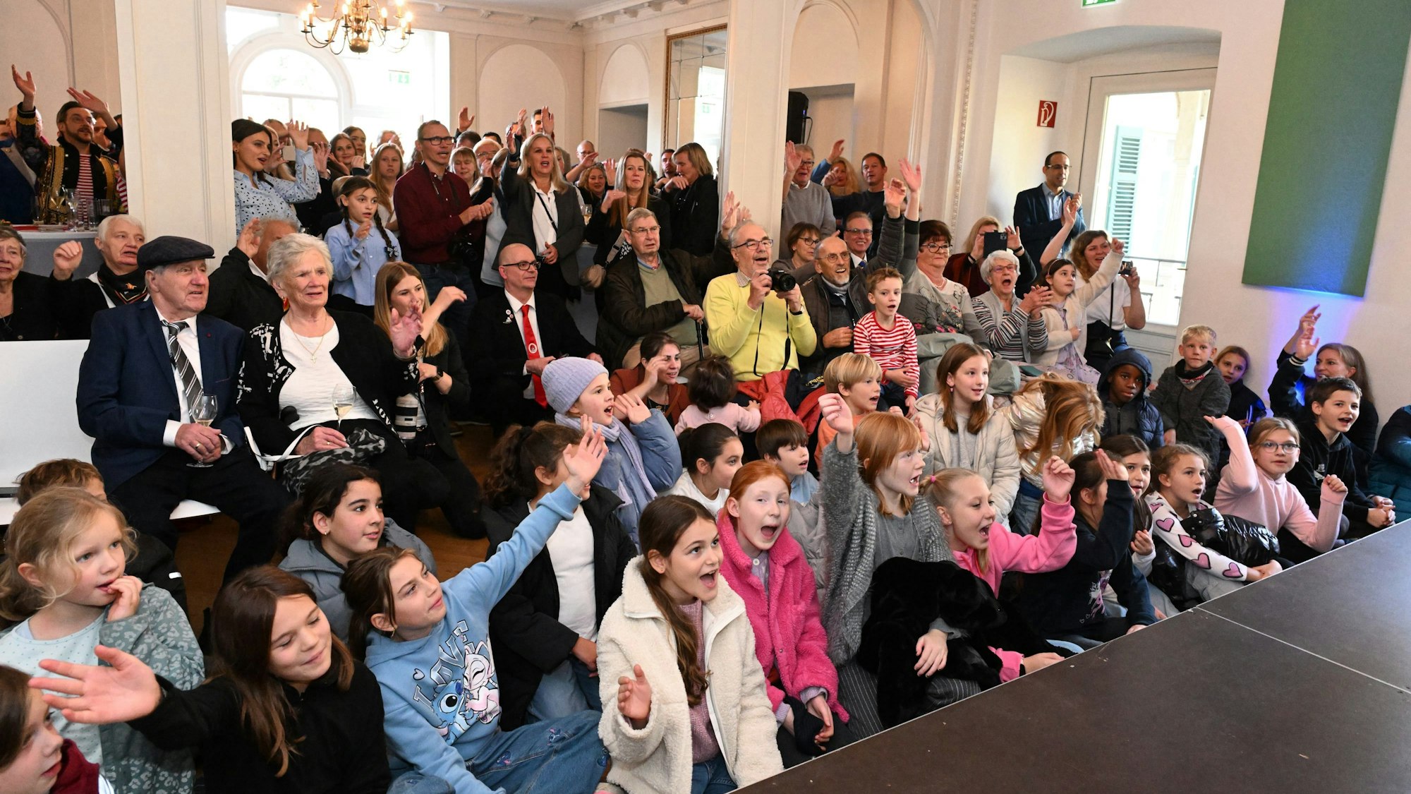 Eine große Gruppe Kinder sitzt vor einer Bühne und gestikulieren zum dreifachen Rösrath Alaaf. Im Hintergrund sieht man Erwachsene stehen die ebenfalls gestikulieren.