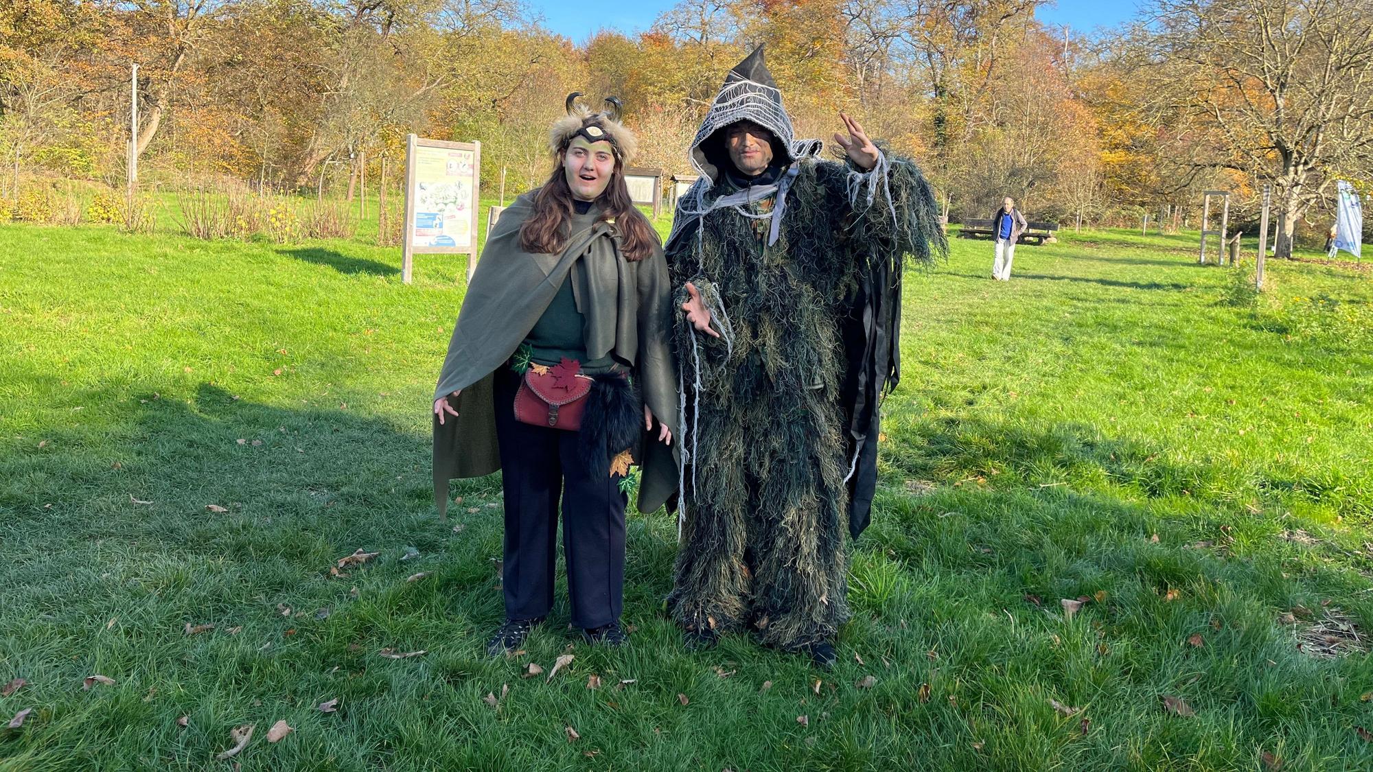 Zwei verkleidete Frauen stehen in Waldgeist-Kostümen auf einer Wiese.