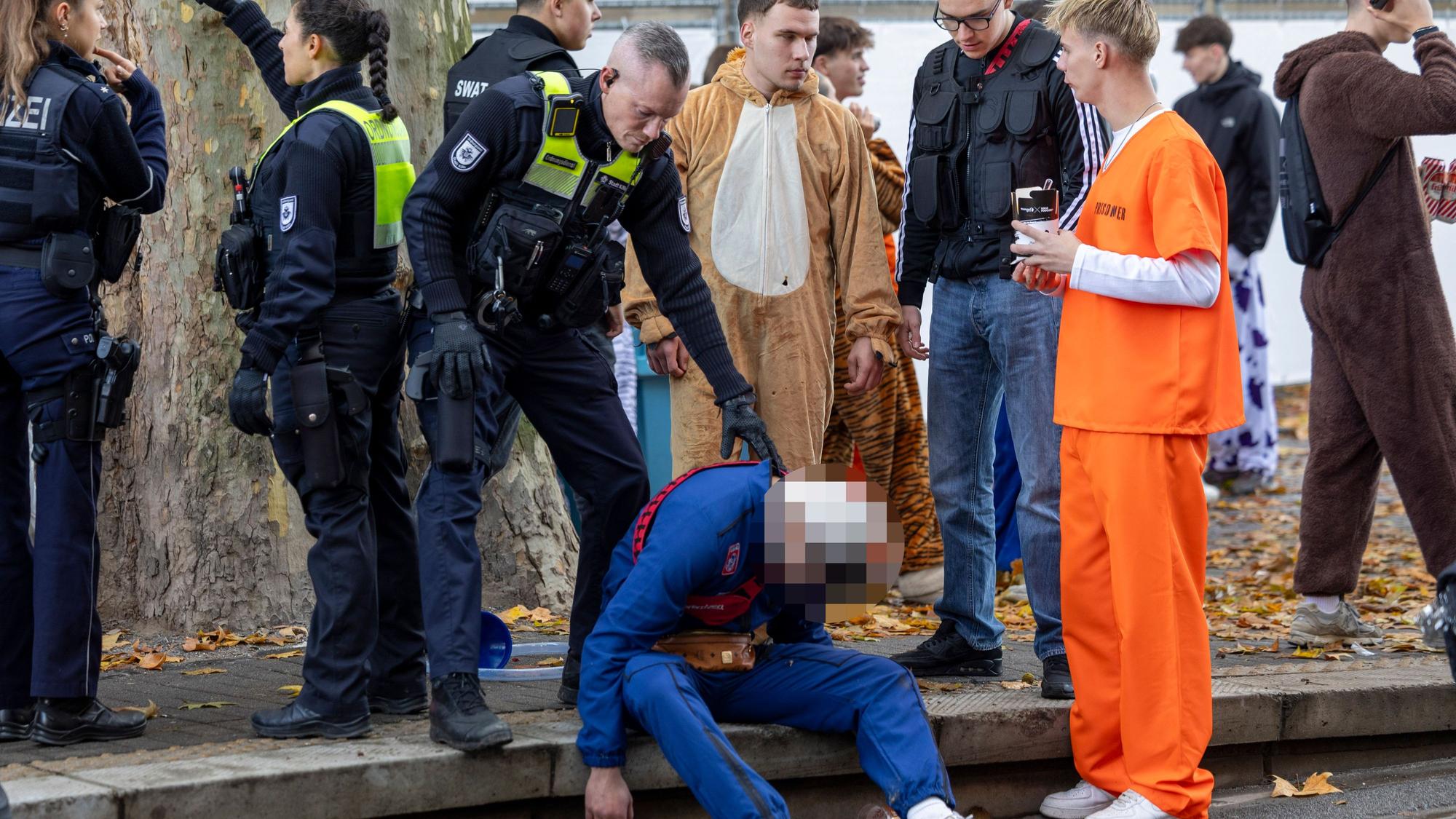 Ein Polizeibeamter und Feiernder stehen bei einem Betrunkenen, der auf dem Boden sitzt.