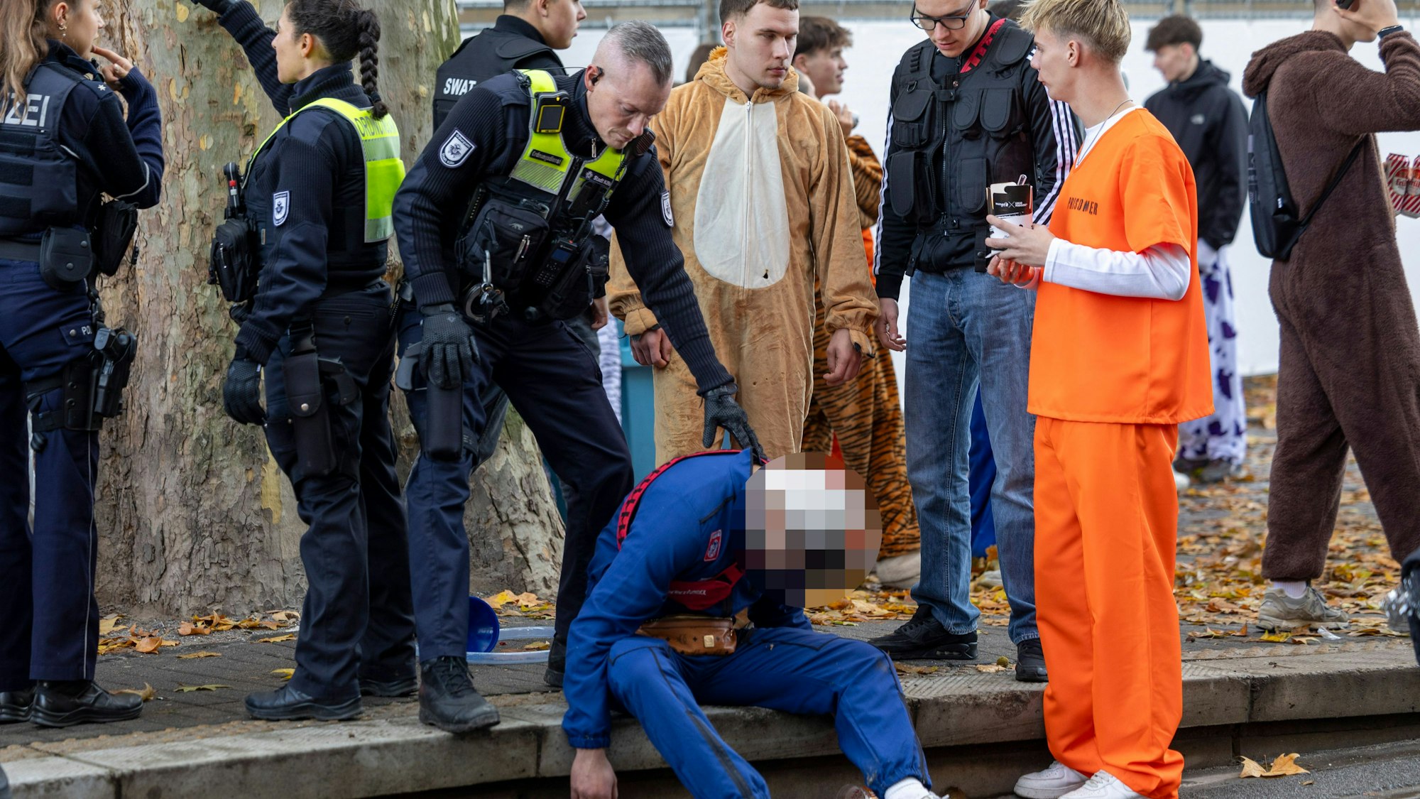 Ein Polizeibeamter und Feiernder stehen bei einem Betrunkenen, der auf dem Boden sitzt.