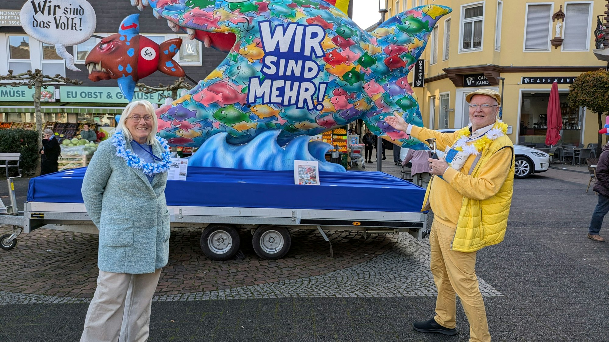 Andrea und Jo Johänning stehen vor dem Wagen, der einen großen Fisch mit der Aufschrift „Wir sind mehr“ darstellt, der such wiederum einen kleinen blau, rot und braunen Fisch schnappt.