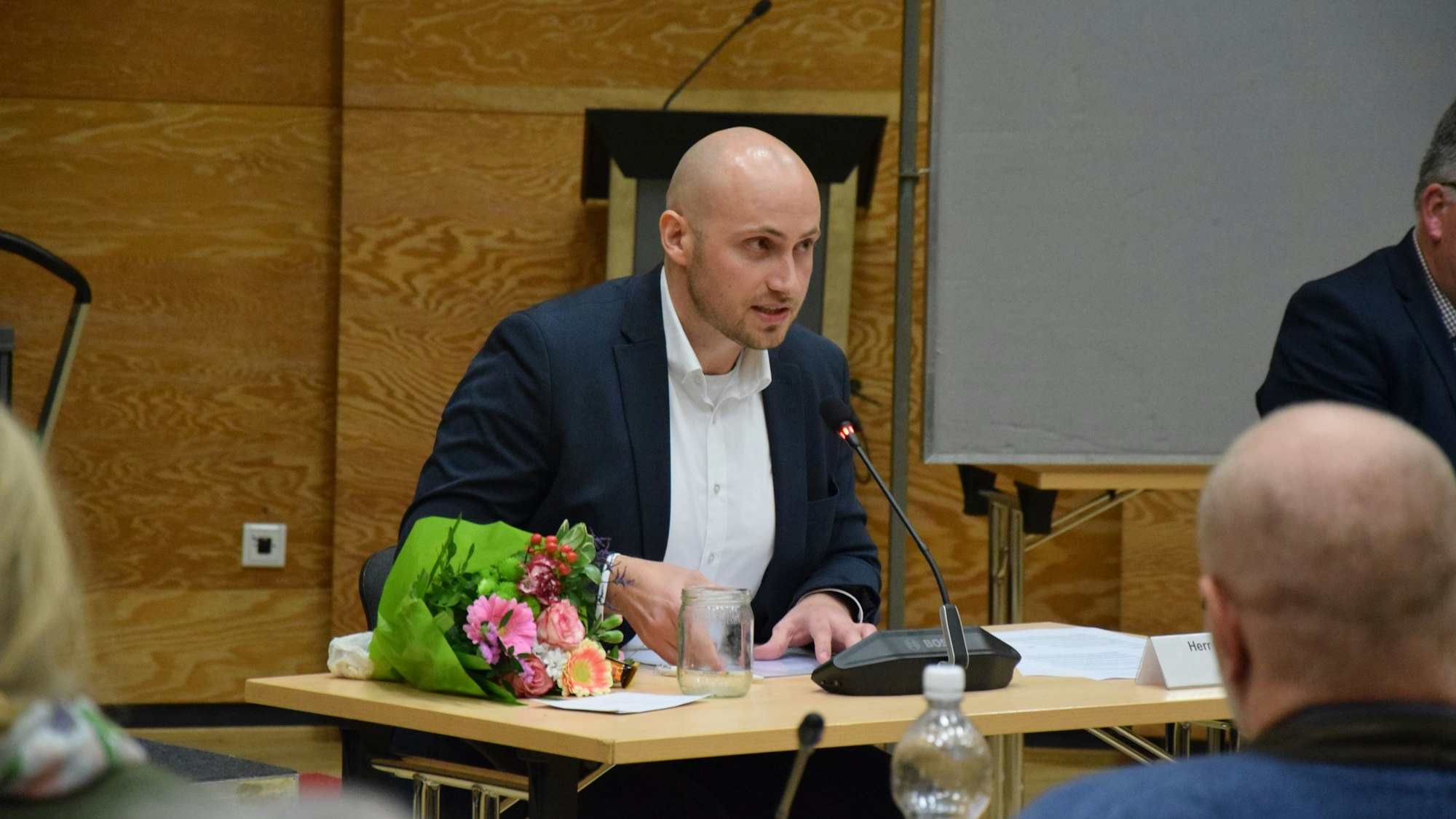 Ein junger Mann mit Glatze sitzt an einem Schreibtisch und spricht in ein Mikrofon.