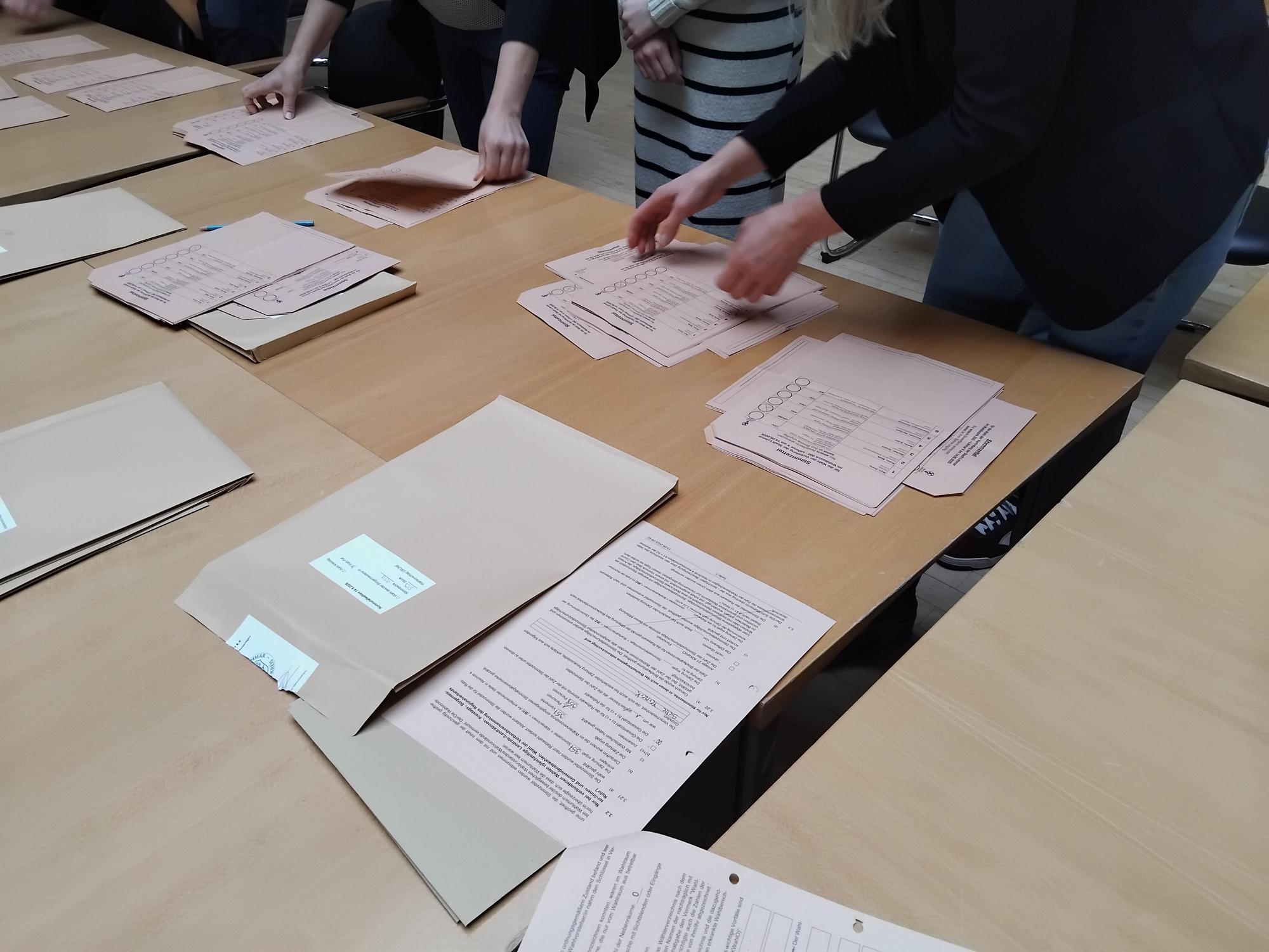 Auf dem Tisch liegen brauen Umschläge und rosafarbene Wahlzettel.