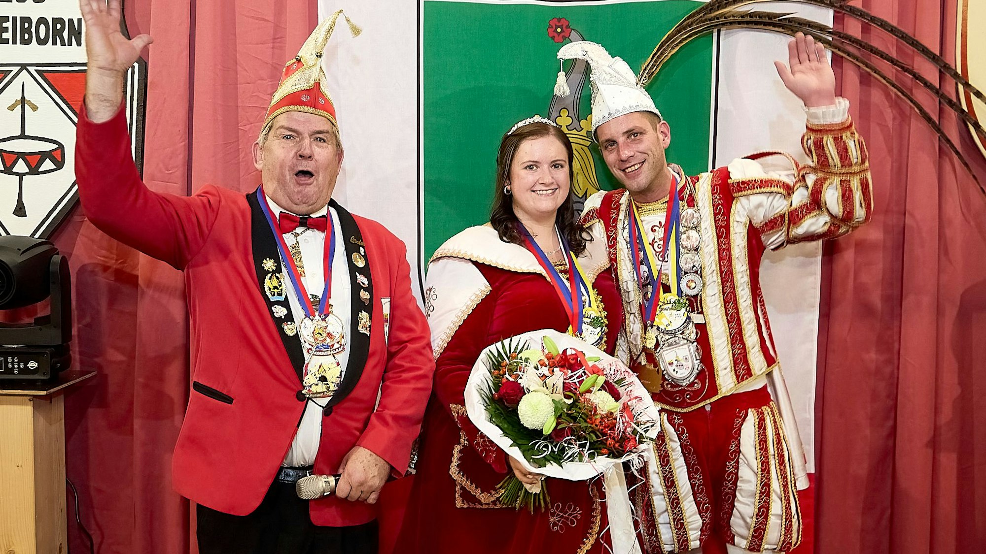Vor einem grün-rot-weißen Vorhang stehen das Prinzenpaar aus Dreiborn im Ornat und der Sitzungspräsident. Die Männer winken.