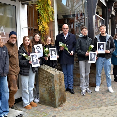 Am Gedenkstein der einstigen Synagoge in Bad Münstereifel stehen einige Menschen. Manche halten Schwarz-Weiß-Fotos oder weiße Rosen in den Händen.