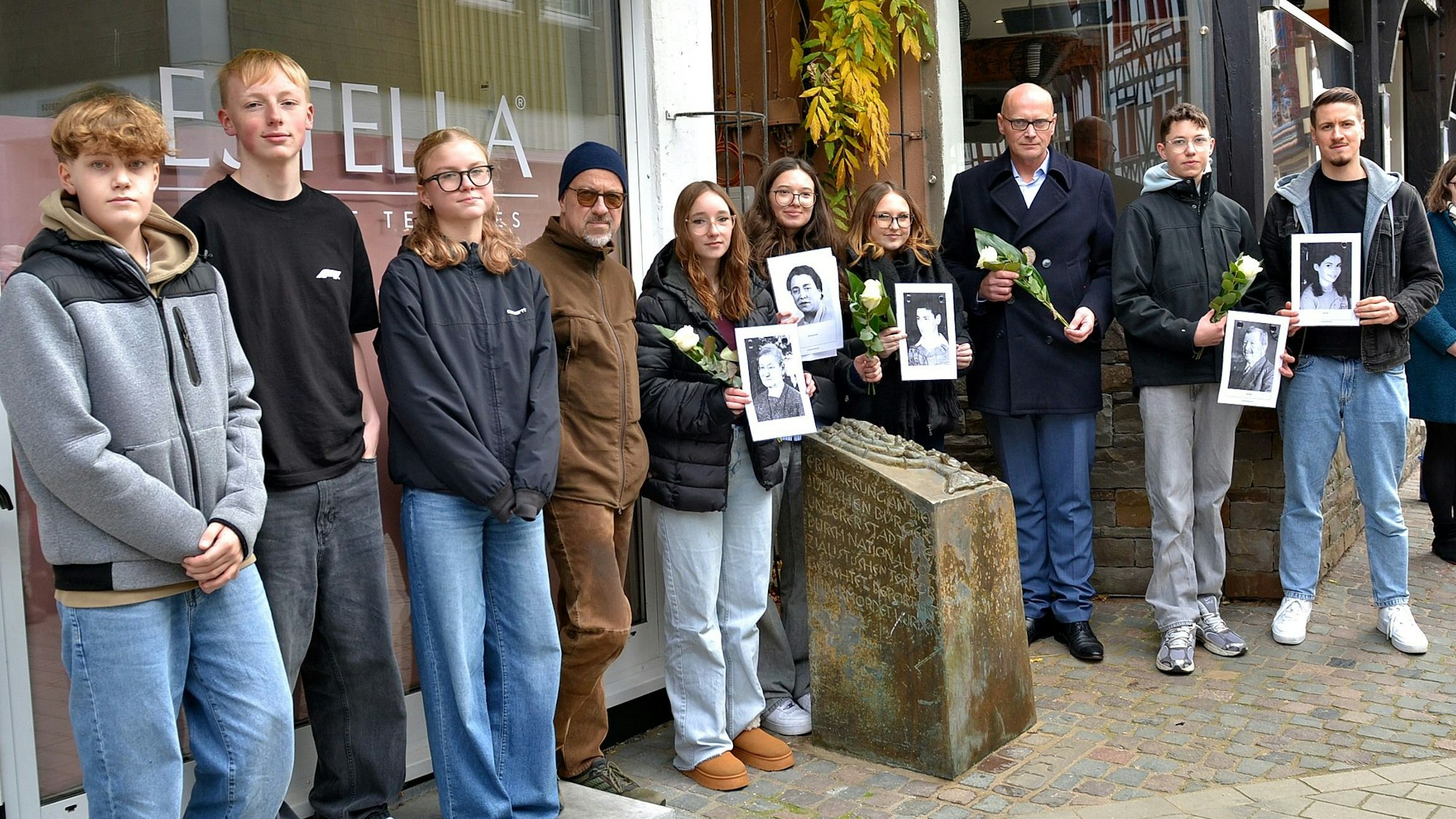 Am Gedenkstein der einstigen Synagoge in Bad Münstereifel stehen einige Menschen. Manche halten Schwarz-Weiß-Fotos oder weiße Rosen in den Händen.
