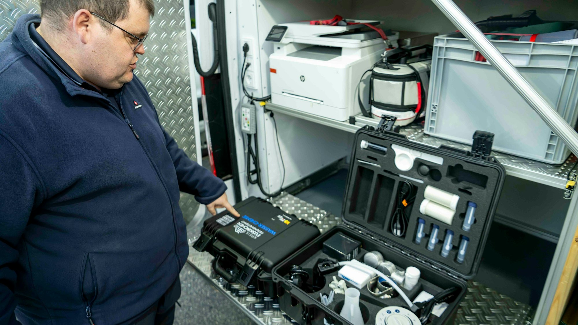 Ein Mann in einer blauen Jacke steht an der geöffneten Hecktür eines Fahrzeugs, in dem mehrere Kisten verlastet sind. Ein Koffer mit technischer Ausrüstung ist geöffnet.