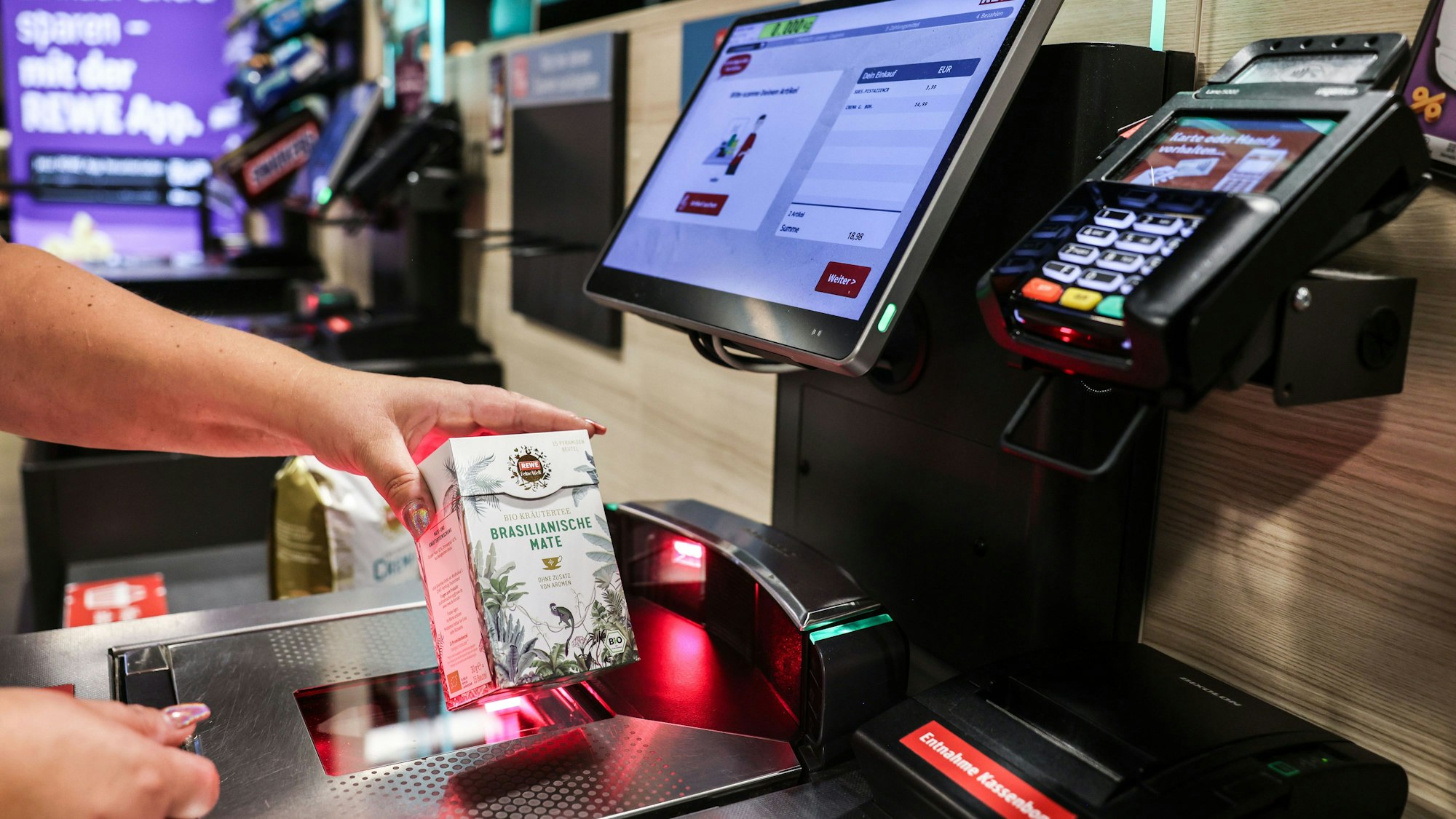 Eine Frau scannt in einer Rewe-Fliale ein Produkt an einer Selbstbedienungskasse.
