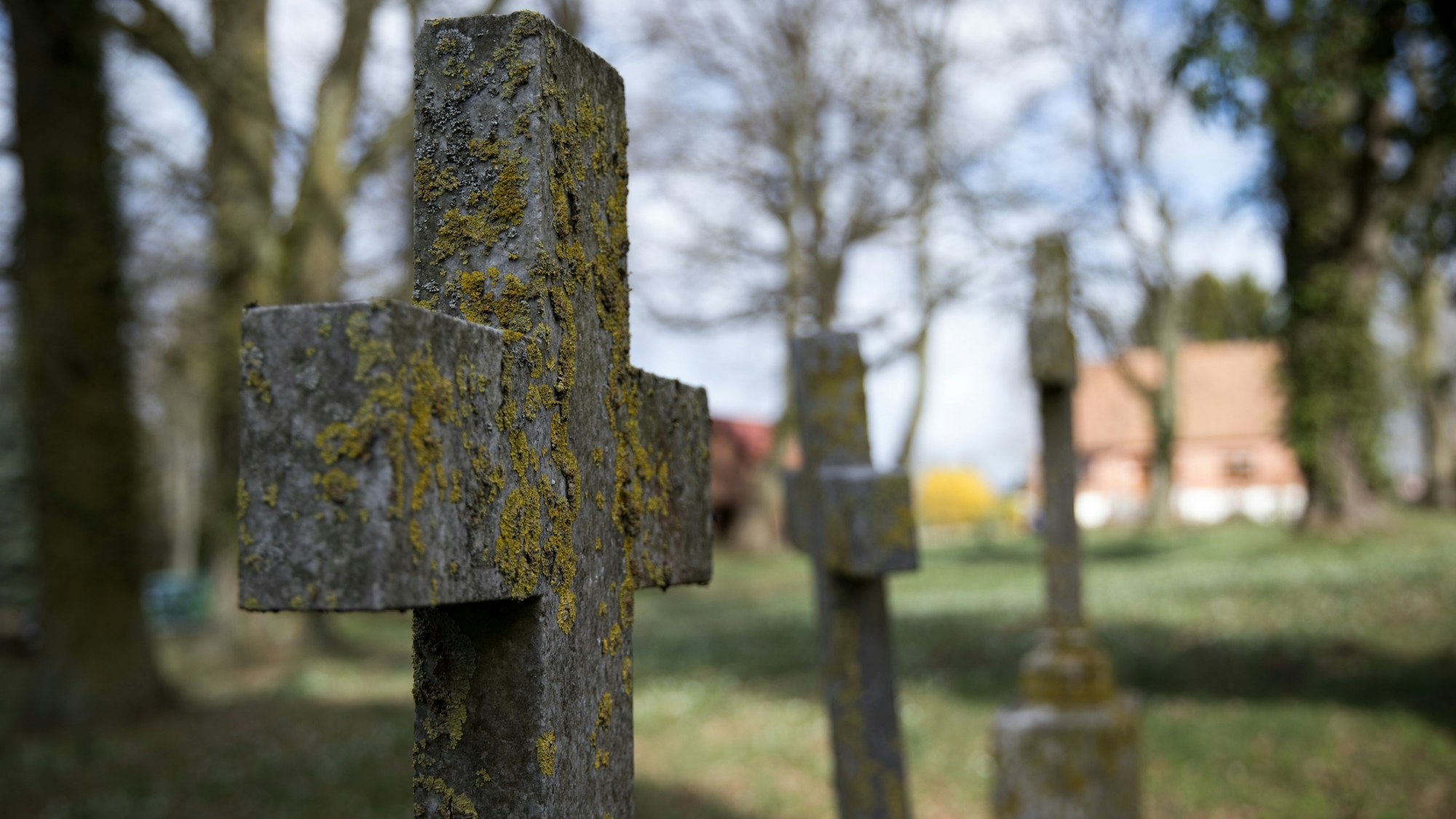 Verwitterte Grabsteine und Kreuze aus Stein stehen vor einer Dorfkirche.