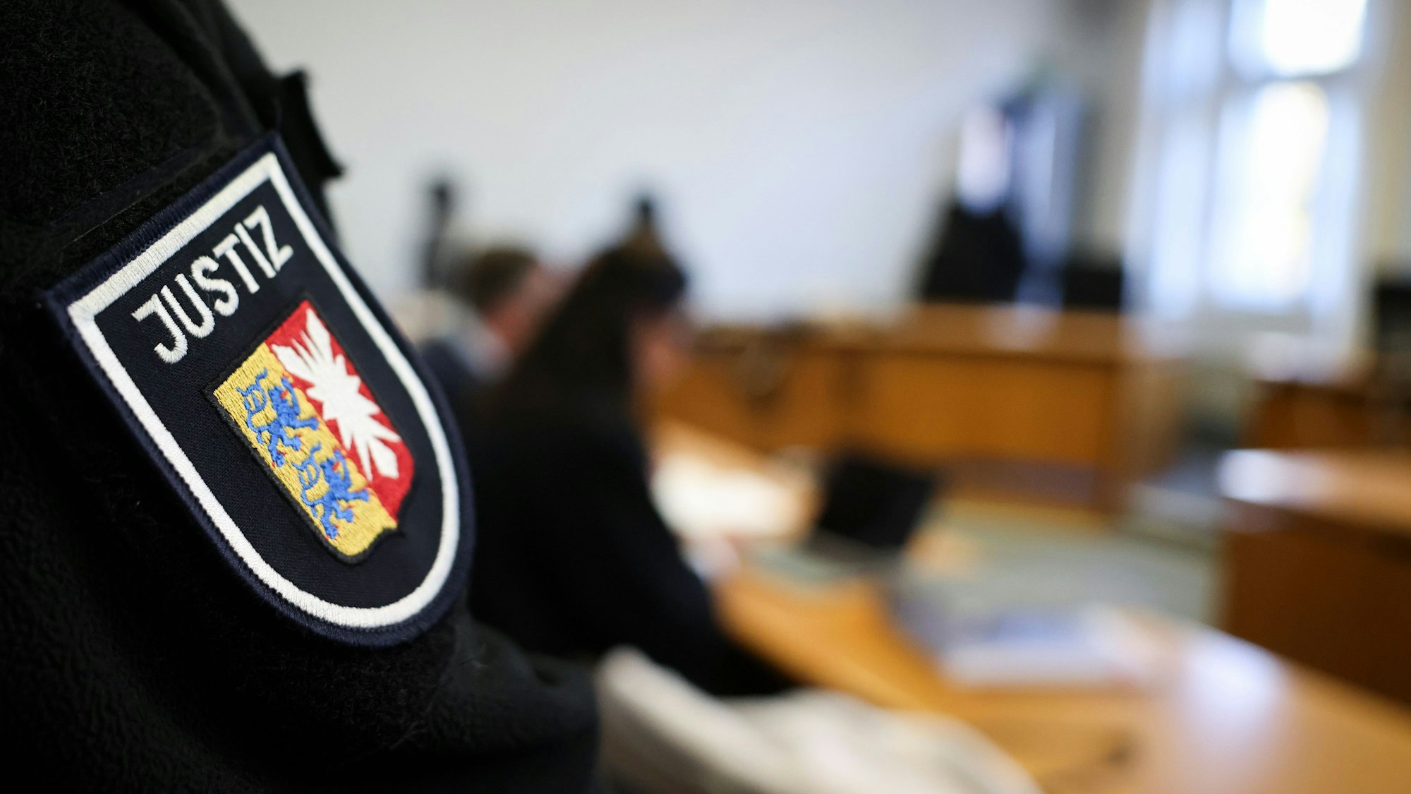 Im Vordergrund ein Arm, auf dem sich ein Abzeichen mit dem Schriftzug „Justiz“ befindet, im Hintergrund ein Gerichtssaal. (Symbolbild)