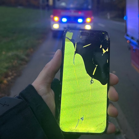 Ein Feuerwehrmann hält ein ramponiertes Handy in der Hand. Im Hintergrund sin Einsatzfahrzeuge mit Blaulicht zu sehen.