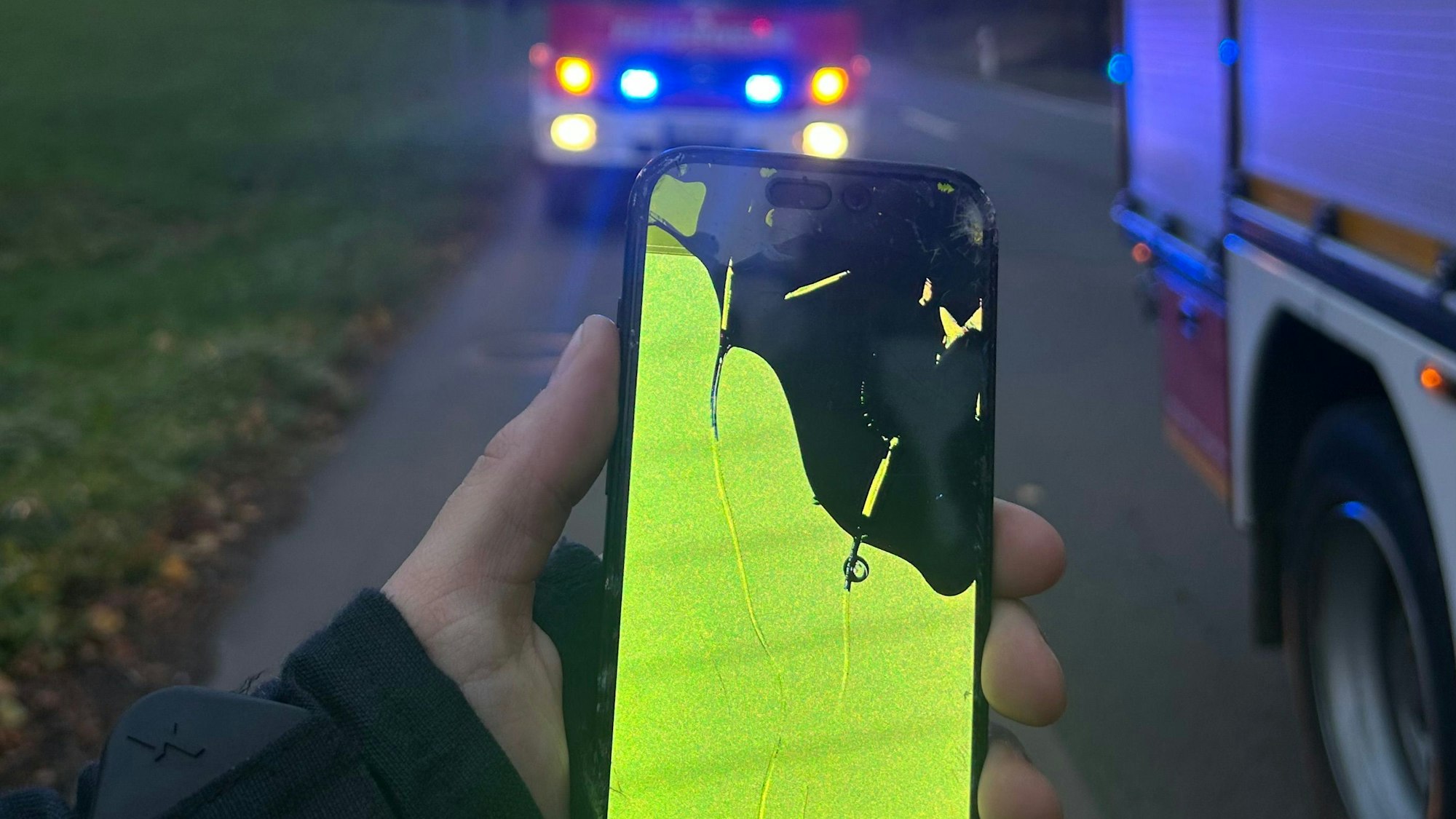 Ein Feuerwehrmann hält ein ramponiertes Handy in der Hand. Im Hintergrund sin Einsatzfahrzeuge mit Blaulicht zu sehen.