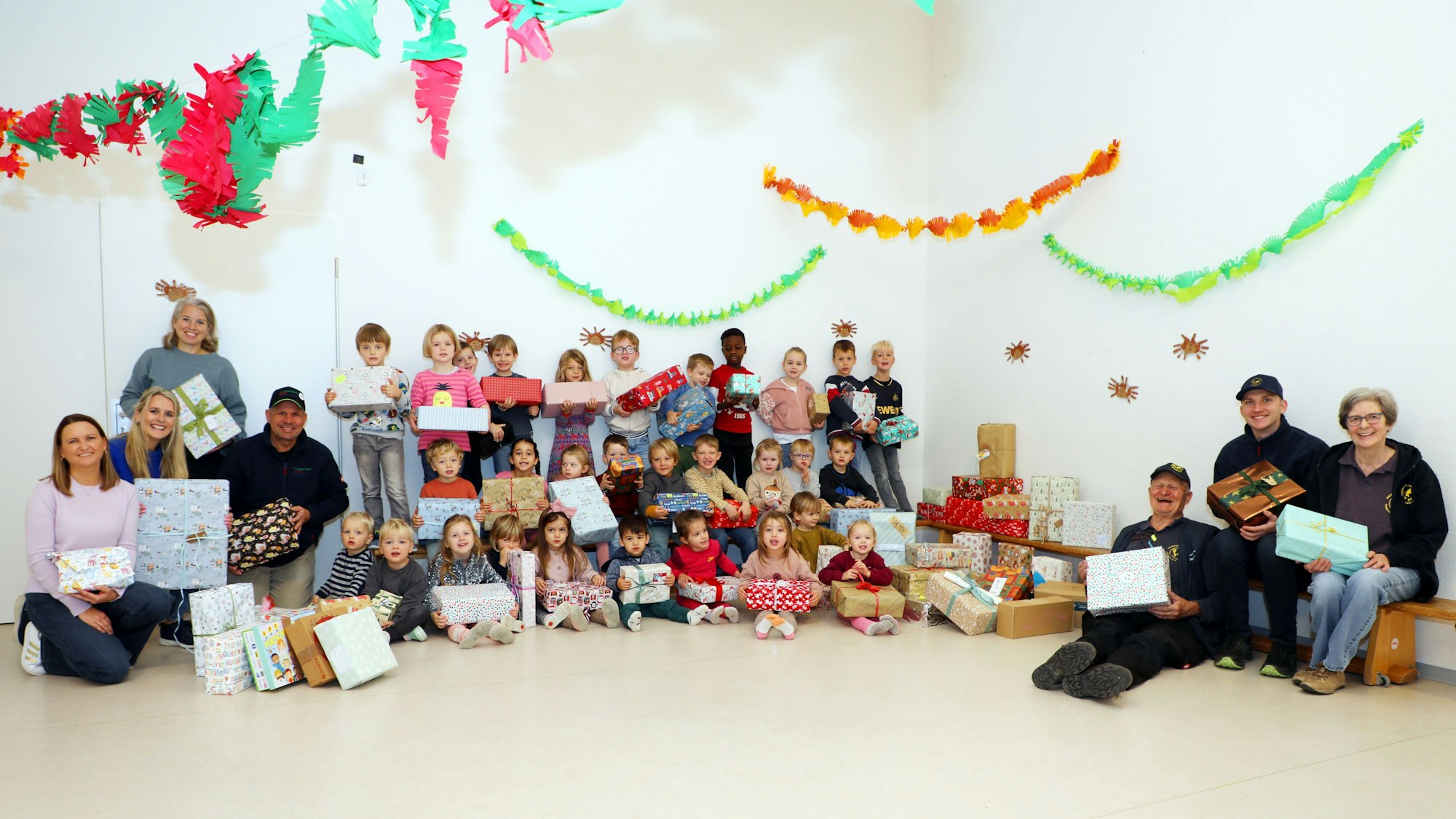 Eine große Gruppe an Kita-Kindern sitzt mit verpackten Geschenken auf dem Boden, links und recht von ihnen sitzen auch Erwachsene.
