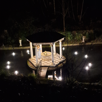 Ein Teich im Dunkeln. Lichter beleuchten eine kleine Insel mit einem Pavillon. Hier sitzt auf dem Brunnenrand der Froschkönig, daneben eine Figur, die aussieht wie eine Prinzessin