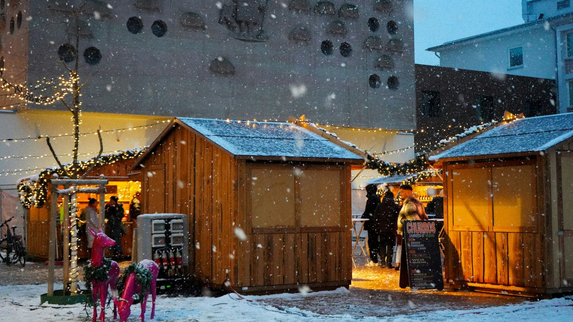 Hütten stehen auf einem Platz, auf dem Boden liegt ein wenig Schnee und es schneit.