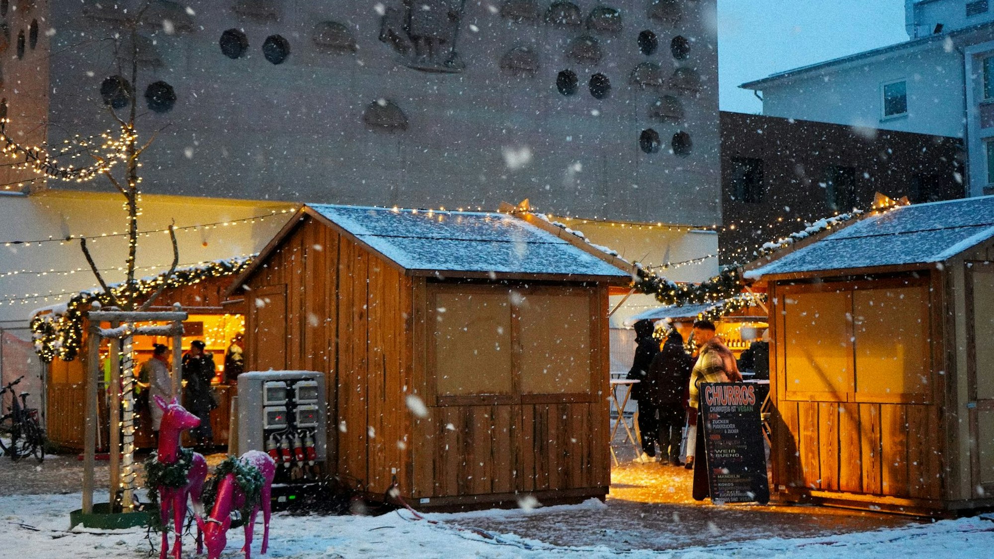 Hütten stehen auf einem Platz, auf dem Boden liegt ein wenig Schnee und es schneit.