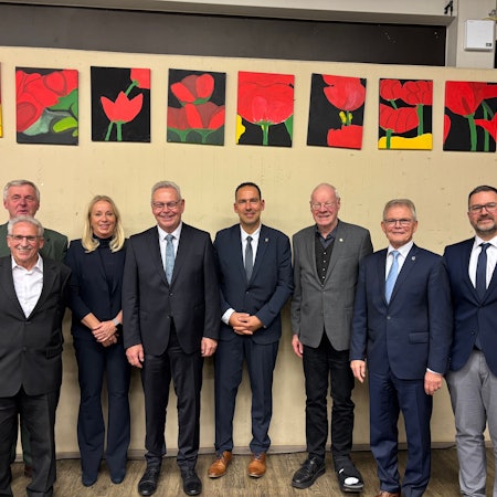 Sieben Männer in Anzügen und eine Frau im Anzug stehen nebeneinander. Über ihnen hängen Bilder von Blumen.