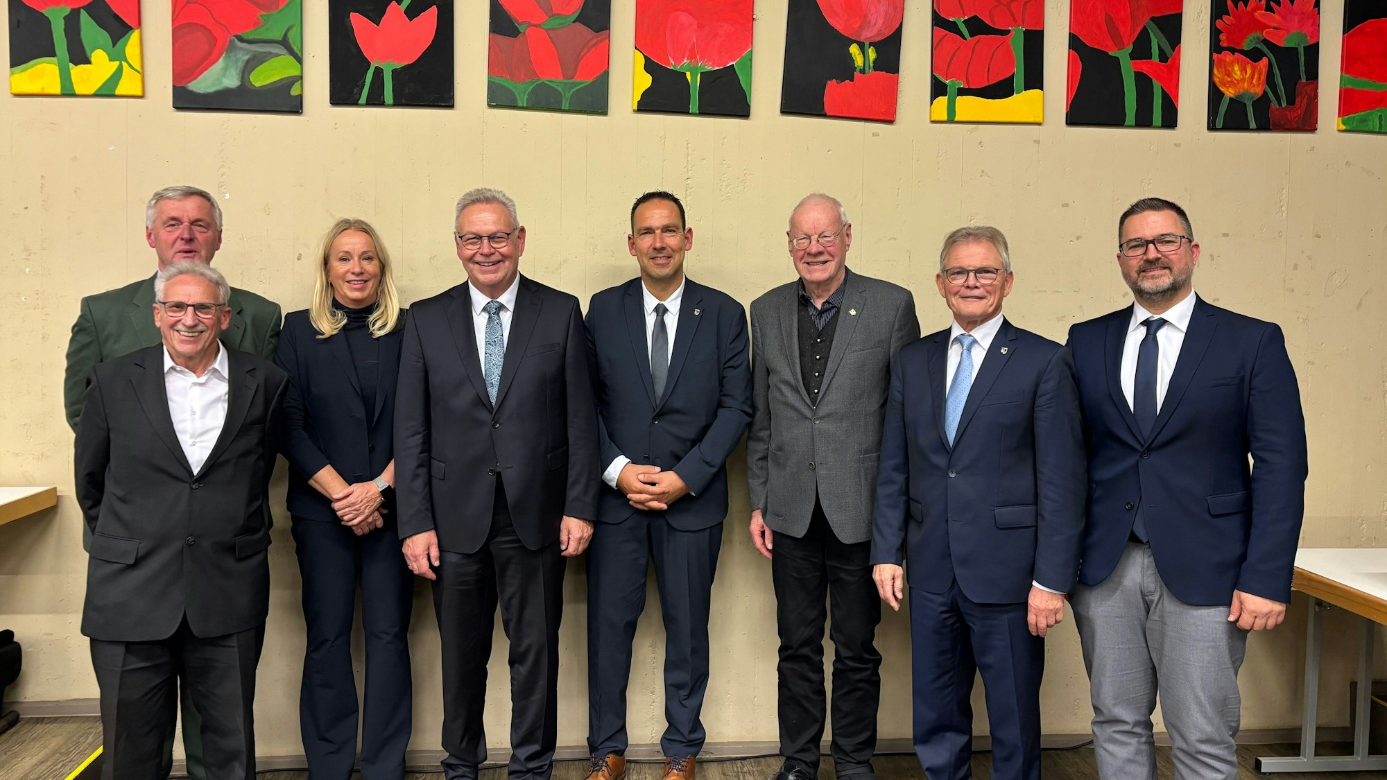 Sieben Männer in Anzügen und eine Frau im Anzug stehen nebeneinander. Über ihnen hängen Bilder von Blumen.