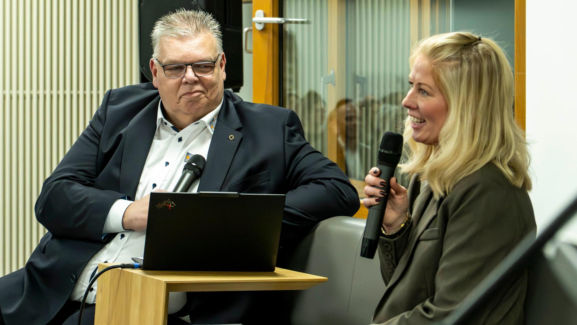 Jochen Kupp und Sarah Komp sitzen auf einer Bank und halten Mikrofone in den Händen.