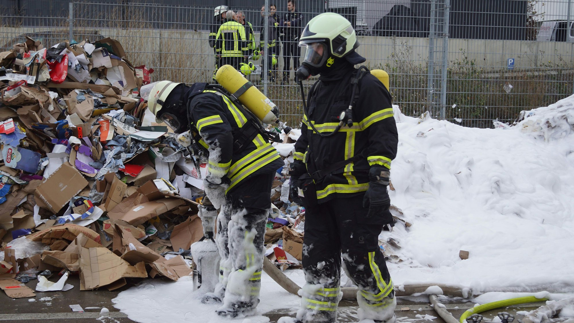 Feuerwehrleute mit Atemschutzgeräten begutachten die abgekippten Verpackungen. Ein Teil davon ist mit Löschschaum bedeckt.