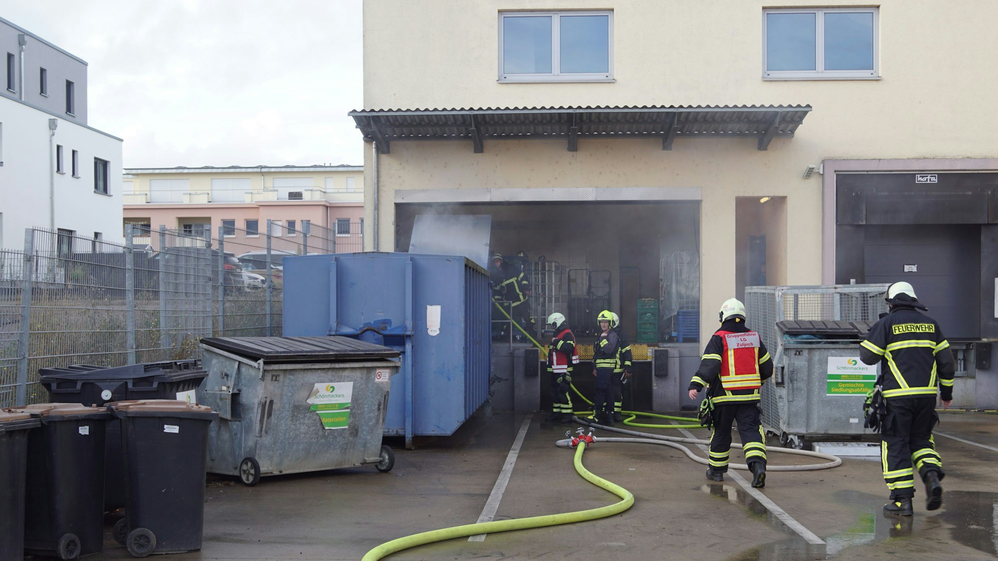 Feuerwehrleute halten ein Strahlrohr in die Öffnung eines blauen Containers, der sich direkt hinter dem Gebäude befindet.