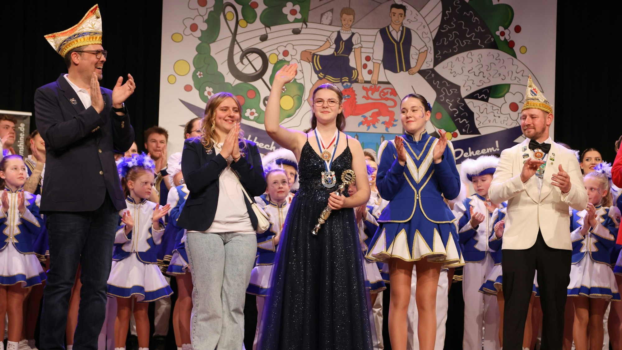 Eine Karnevalsprinzessin steht samt Gefolge auf einer Bühne und winkt ins Publikum.