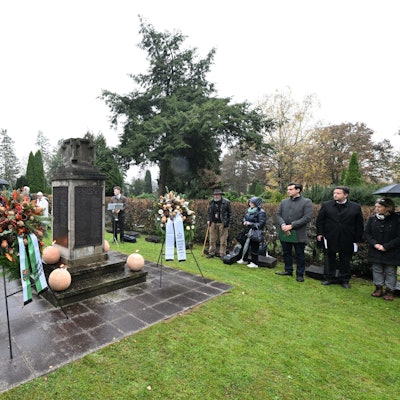 Eine Trauergemeinde hat sich um ein Ehrenmal mit Trauerkränzen versammelt. Die Besucher tragen dunkle Kleidung, einige halten Liedtexte in der Hand.