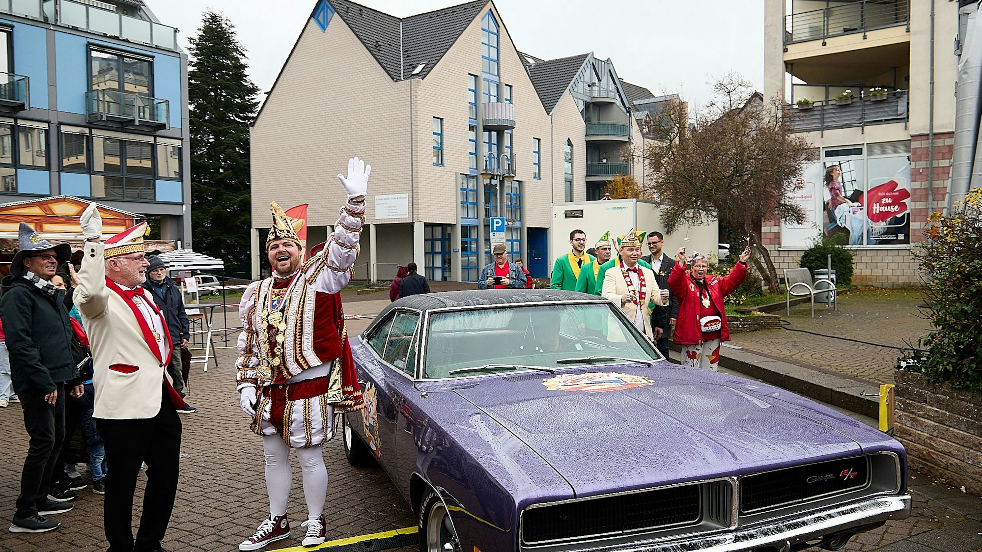 Der Prinz steht im Ornat vor dem violetten amerikanischen Schlitten und reckt die Hand zum Jubel hoch.