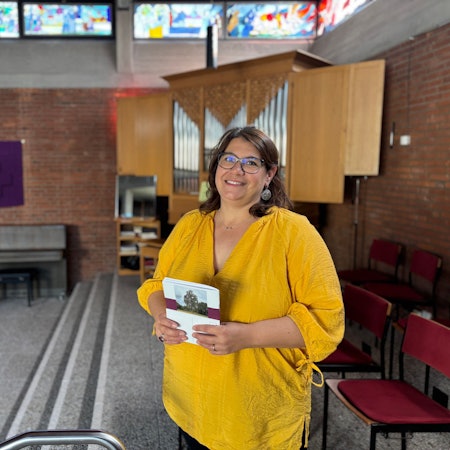 Judith Weichsel hält die Zeitung der Evangelischen Kirchengemeinde Weilerswist vor sich. Sie steht in einem Raum der Martin-Luther-Kirche, in dem sich eine Orgel befindet.