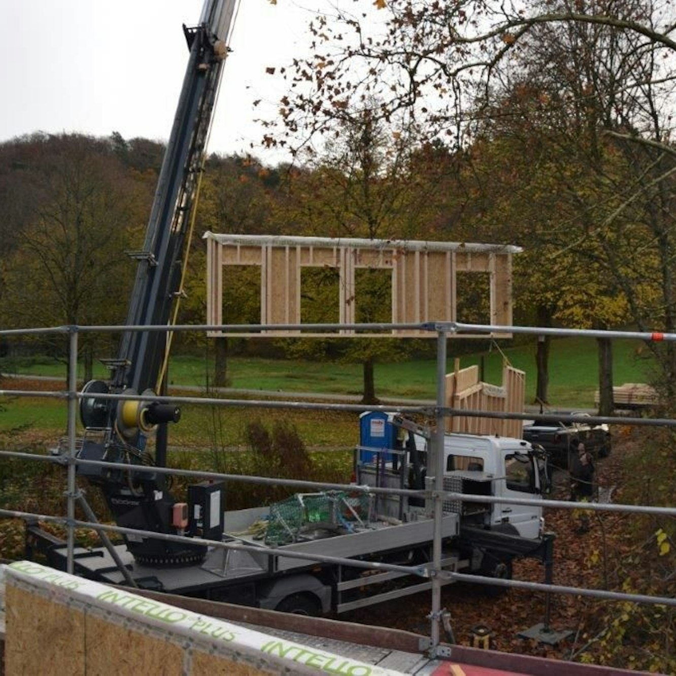 Ein Kran hat ein hölzernes Fassadenbauteil mit vier Türöffnungen am Haken.