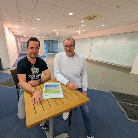 Das Foto zeigt die beiden Betreiber auf Hockern an einem Stehtisch in den Räumen der früheren Postfiliale im Hürth-Park.