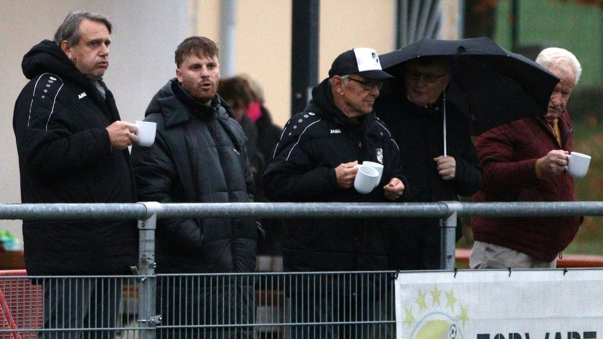 Fünf Männer beobachten das Spiel. Drei von ihnen haben Tassen in der Hand, einer sogar zwei. Einer der Männer trägt einen geöffneten Regenschirm. Der Mann ganz links bläst die Backen auf.